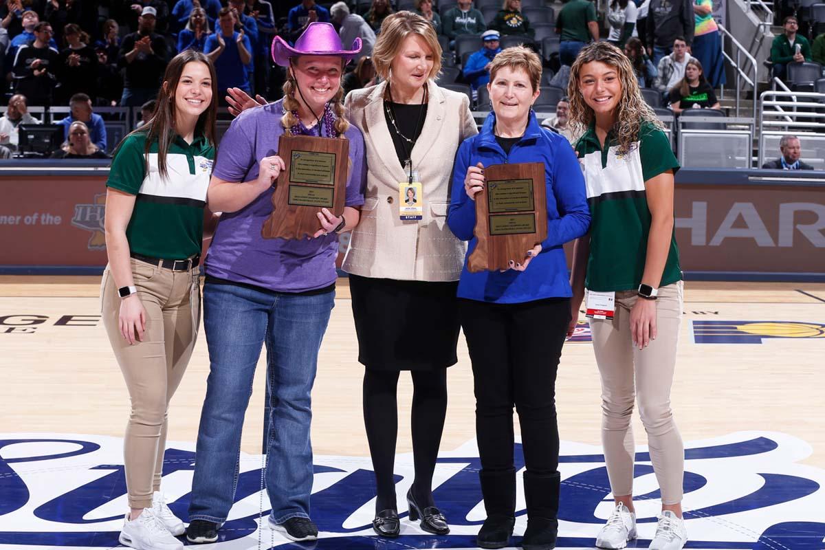 Students and teachers pose with the IHSAA assistant commissioner at center court of the Class 1A girls basketball state finals for the inaugural IHSAA Champion of Education Award presentation.